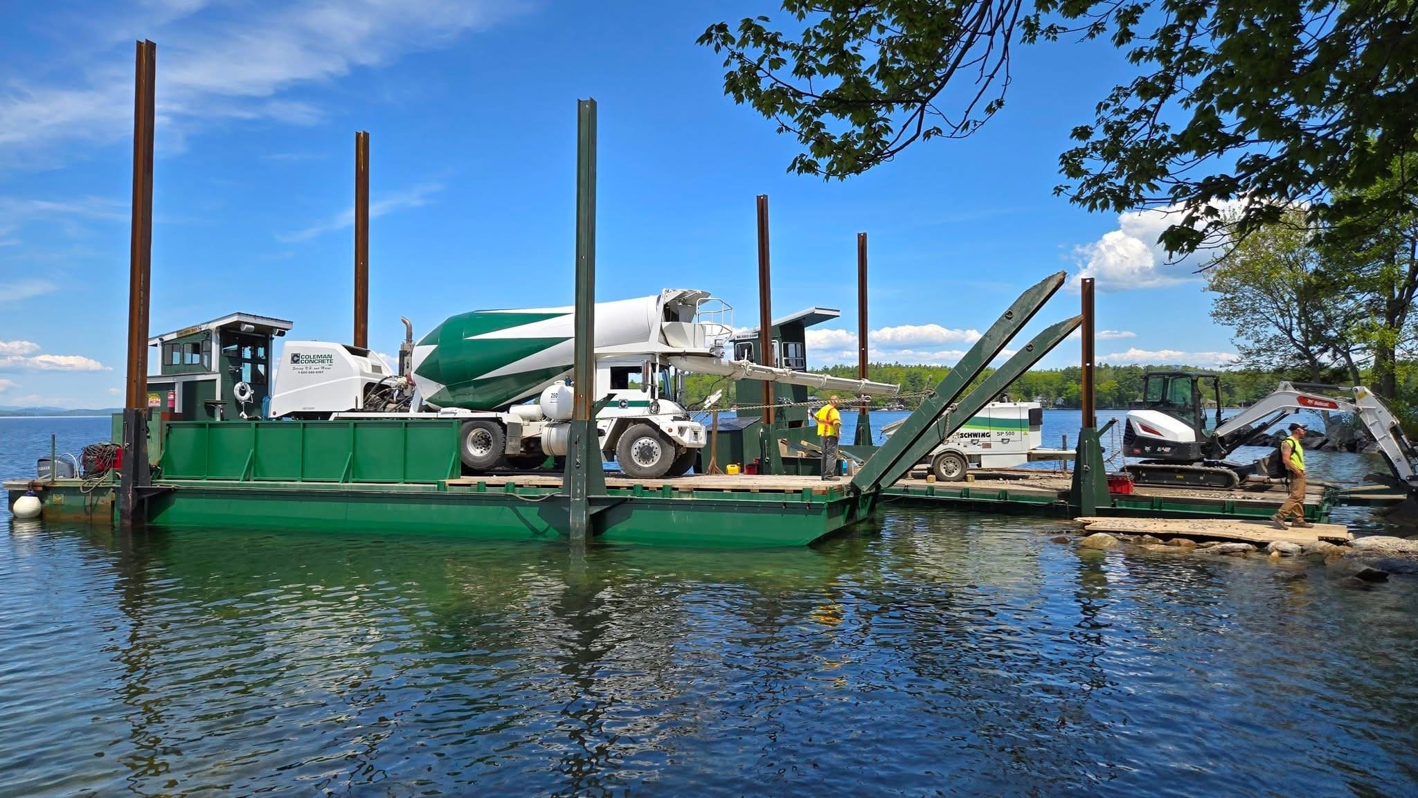 Concrete mixer truck loaded on a green barge under summer foliage