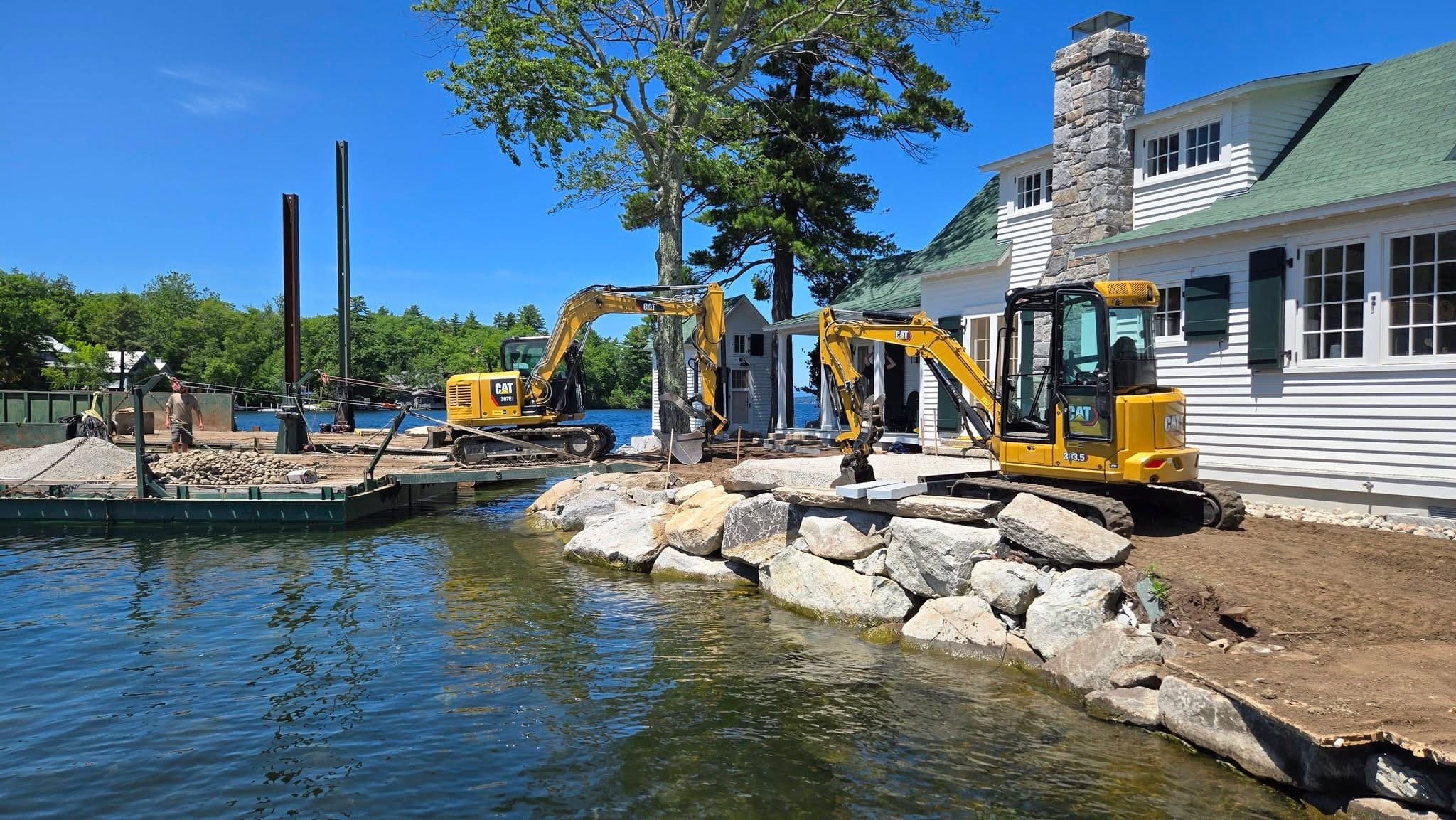 Two excavators and retaining wall under construction at a white-clapboard home