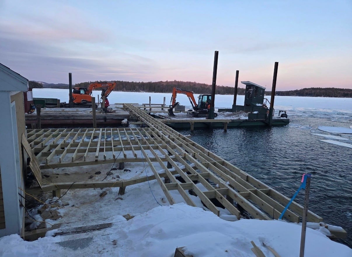 Wide framing of dock joists with two excavators in the distance and a pink dawn sky
